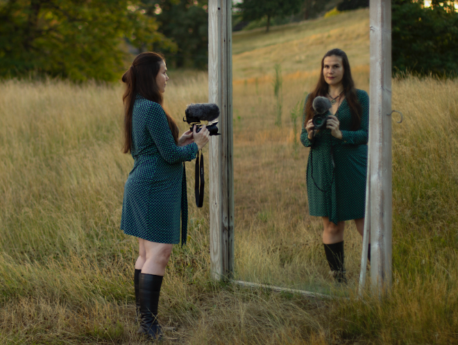 Hidalgo stands before a large mirror in a field with her video camera. As she looks into the mirror, we see her reflection looking back at us.