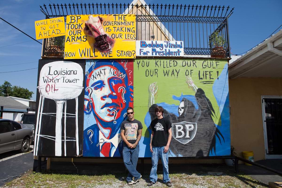 A color photograph depicting Bobby Pitre and his assistant on US Highway 1 in front of the Obama What Now? mural made in response to the BP oil spill and lack of
          governmental action to address local needs.