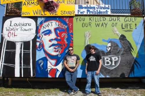 Two young men standing in a front of a wall with graffiti. A grafitti Obama Hope with the words What Now and question marks is visible to their left.