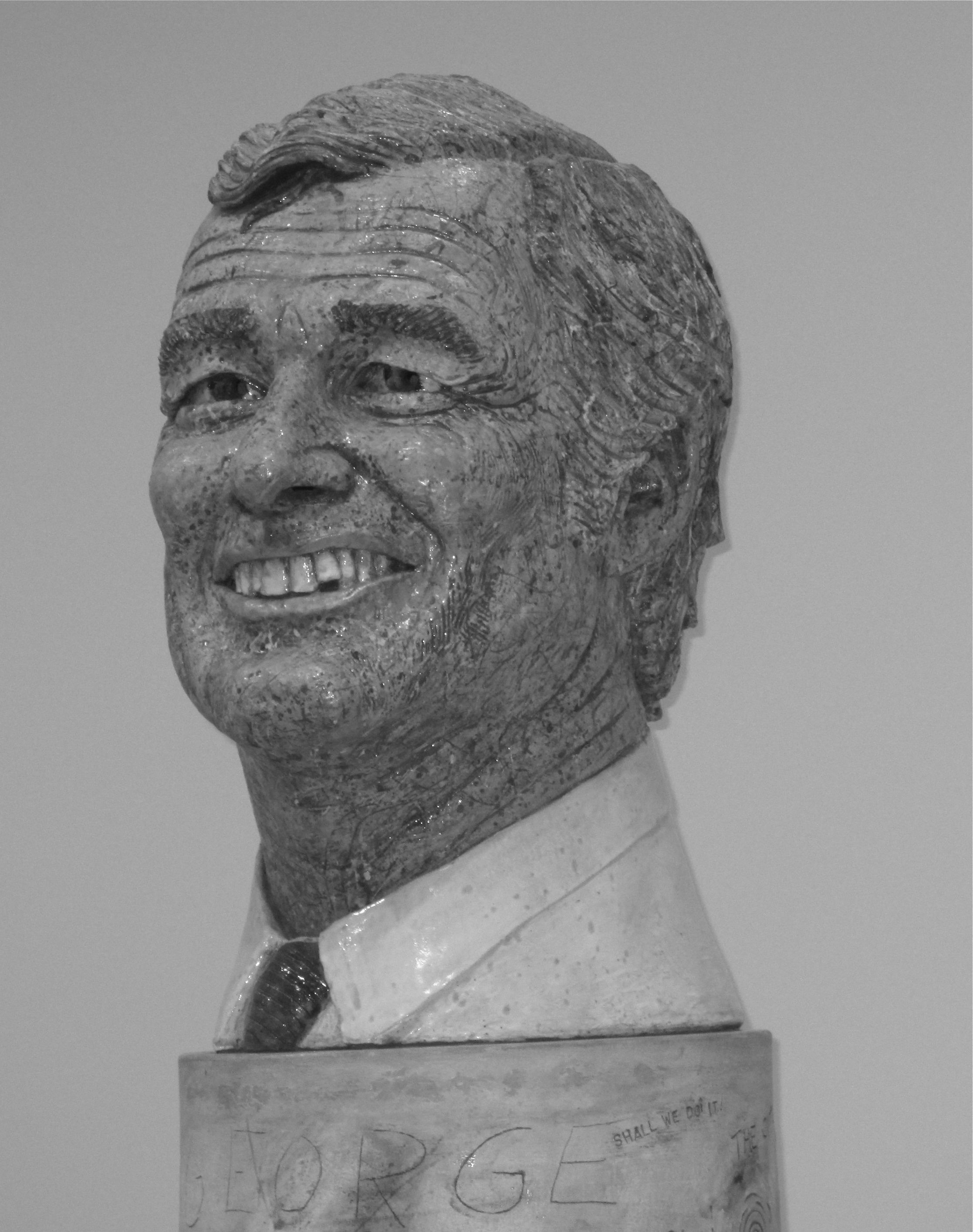Figure 4 This photo is a close-up image of the head of the George Moscone bust. It is extremely photo-realistic, portraying wrinkles, glossy hair, and an unflattering smile. The word “George” is roughly carved under the bust.