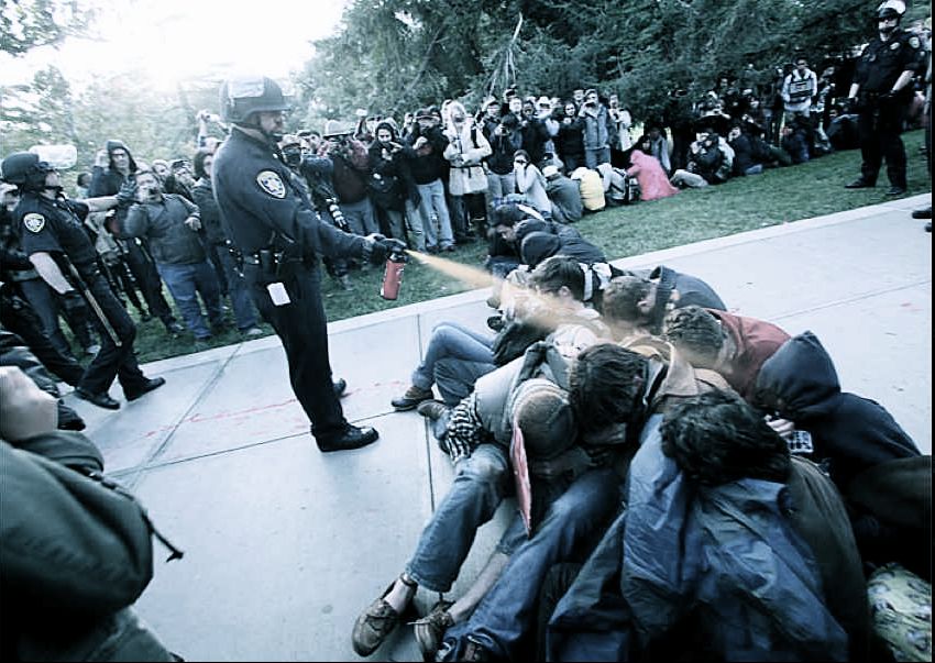 Figure 7 A photograph of Lieutenant John Pike pepper spraying  UC Davis students during an on-campus protest in 2011. Students are seated on the ground as Pike stands above them spraying.