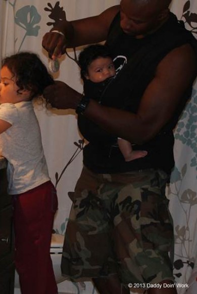 Photo showing Richards wearing his infant daughter in a front baby carrier while brushing his older daughter’s hair