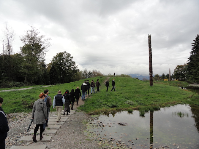 a line of people walking onto a stone path through a grassy park, a totem pole can be seen in the distance