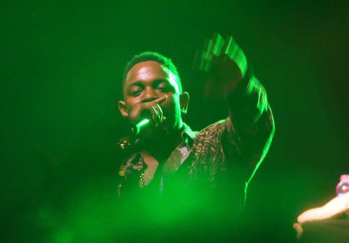 Kendrick Lamar, holding a microphone and holding his left hand out; picture is blurry and cast in green lights