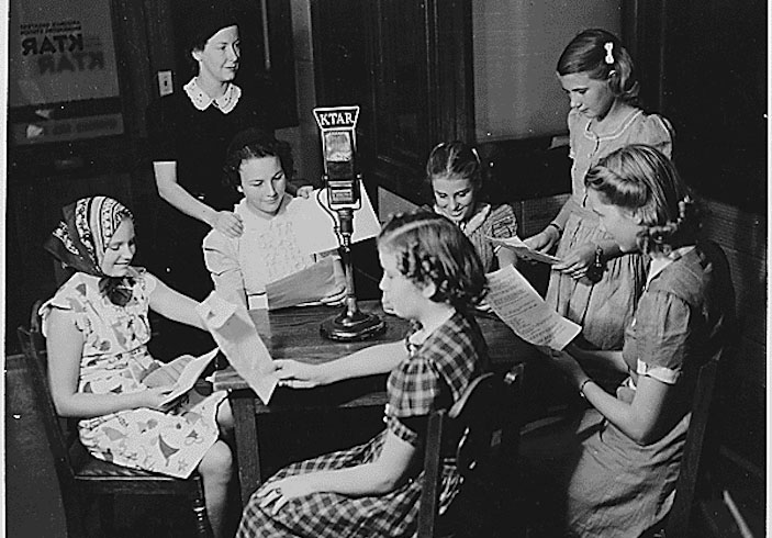 Seven young women sit and stand around a table; each holds onto paper, likely scripts; an old fashioned microphone sits in the center of the table