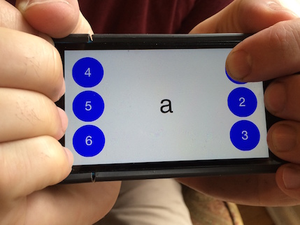 Georgia Tech's BrailleTouch app displayed on a smartphone screen. Hands wrap around the smartphone, touching six large braille dots. The center of the screen diplays u in both the roman letter and braille grid