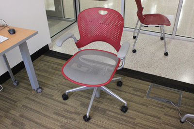 A moveable chair with a grey seat and red back is placed in front of a table on wheels.