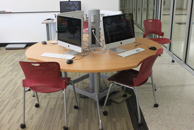 A round table has four computers on it, each one facing out so that the backs of the computers face each other. There are chairs with wheels placed in front of each computer station. 