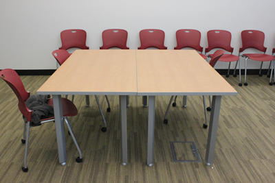 One chair is placed at a large wood-finished square table. Six red chairs line the wall behind the table. 