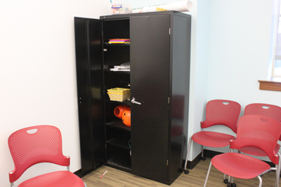 A black storage cabinet is placed in the corner of the room. There are three redish chairs placed next to the cabinet. 