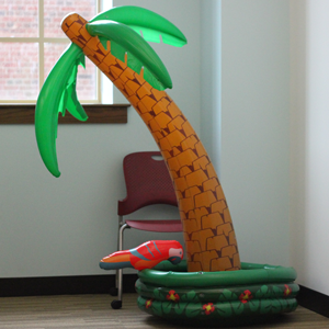 An inflateable palm tree sits in the coner of the room. One chair is placed behind the tree. 