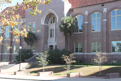 The Johnston Building at FSU is a two story, historical building with large, pane glass windows, brick exterior walls, and an arch entrance. There are stairs to the front entrance and windows below the first floor, suggesting a basement level. 