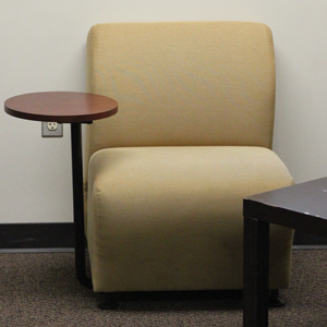 A beige chair sits along a wall. The chair has a small, round table fixed to one of the arm rests. 
