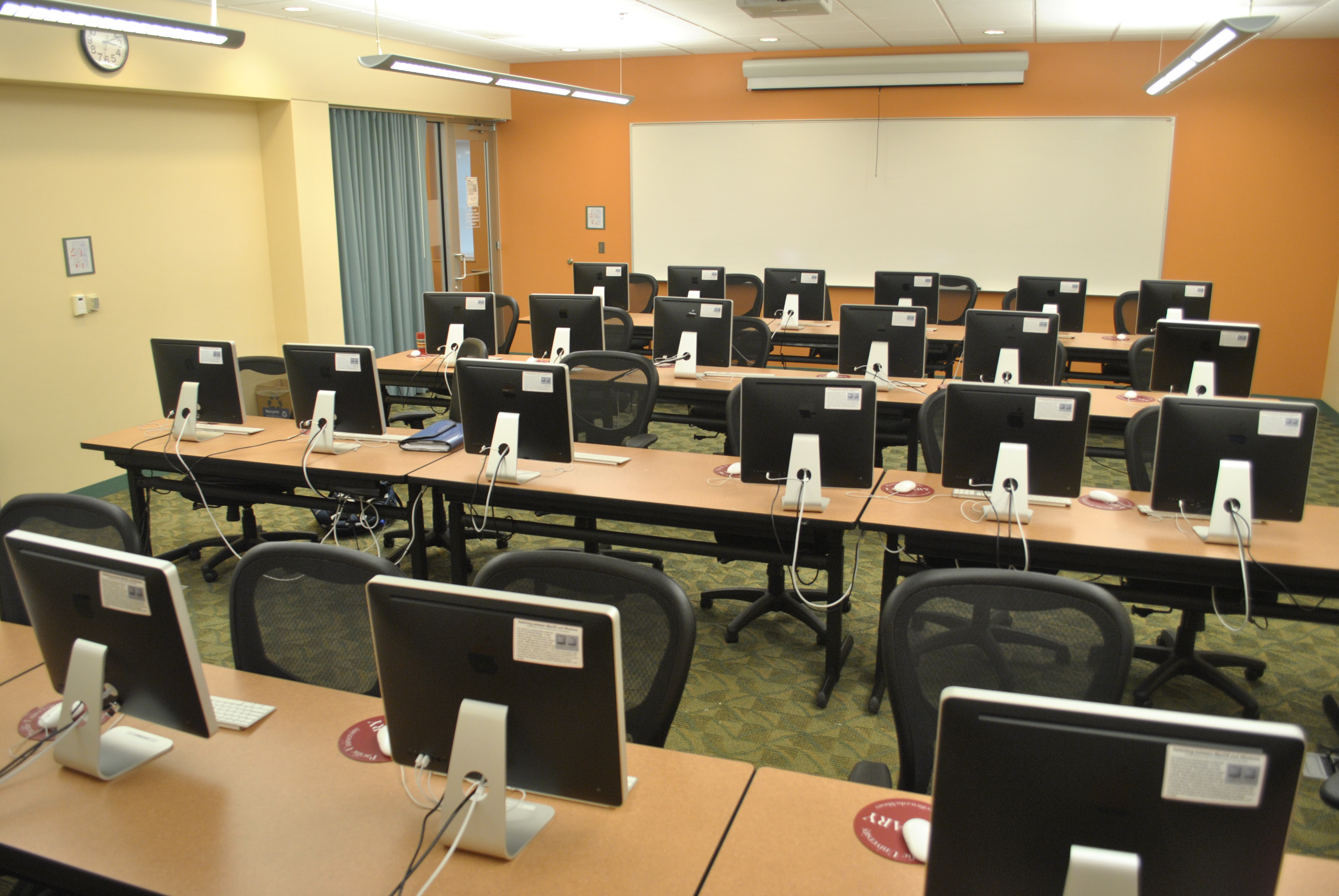 Apple computers are lined up on long desks. There are four rows of tables with monitors. 