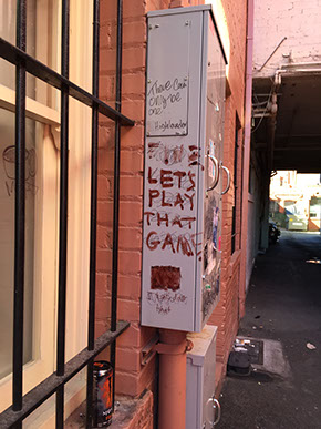 Graffiti: LET'S PLAY THAT GAME painted in red on a gray fusebox.