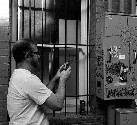 (Black and white photo): Jonathan, standing in front of a window protected by iron bars, focuses his smartphone for another picture.