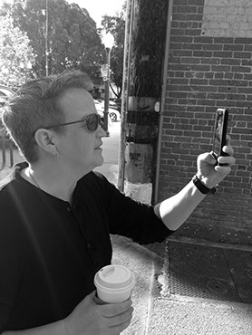 (Black and white photo): Wielding a go-cup of coffee in her right hand and an iPhone in her left, Jackie takes a picture. 