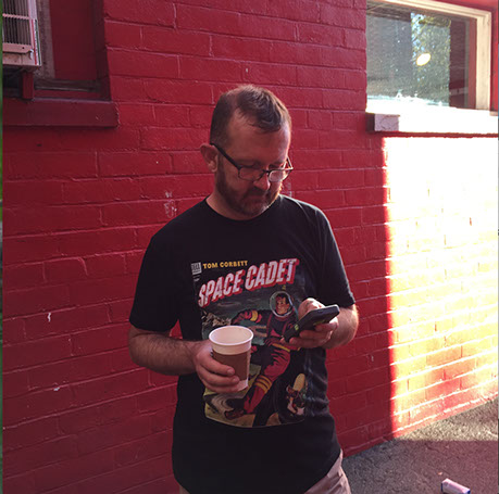 In front of a red brick wall, Jonathan (in a Space Cadet t-shirt) texts while holding a go-cup of coffee in the Augie's alleyway.
