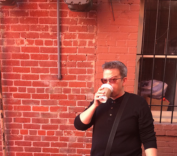 Jackie with sunglasses, drinking coffee in the Augie's alleyway. Behind her is a red brick wall and a window with iron bars over it.