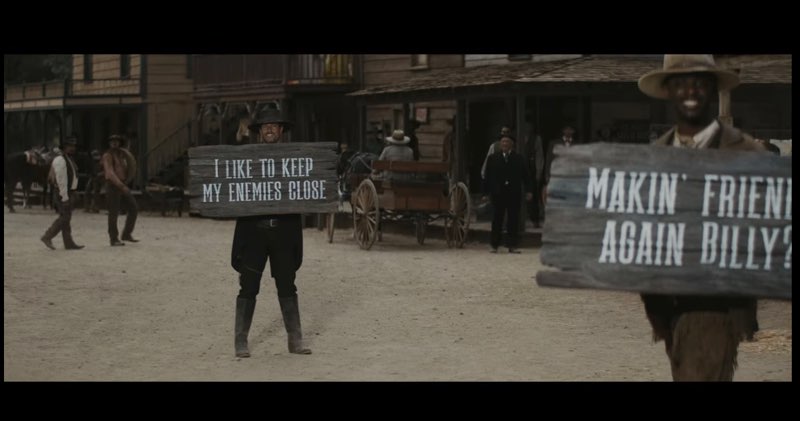 Two cowboys stand outside in an Old West scene and smile at the camera. One cowboy is holding a wooden sign that reads, "I like to keep my enemies close." Another cowboy, played by Monte Bell, is holding a wooden sign that reads, "Makin' friends again Billy?"