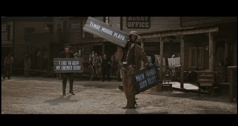 Two cowboys are standing outside in an Old West scene. One cowboy is still holding the sign that reads "I like to keep my enemies close." The cowboy played by Monte Bell has a wooden sign in each hand. The wooden sign in his left hand reads, "Makin' friends again Billy?" The wooden sign in his right hand reads, "Tense music plays."