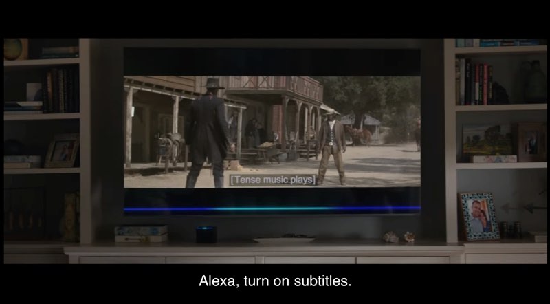 The camera shows a television in a living room. On the television screen, two cowboys are facing each other in an outdoor scene in the Old West. Closed captions on the television screen in this living room read "[Tense music plays]." At the very bottom of this frame are our own closed captions, which read, "Alexa, turn on subtitles."