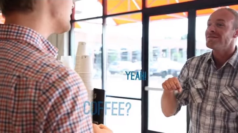 A male barista and a male customer are facing each other. The male customer is signing with the large blue subtitles showing, “YEAH!” next to his signing hand. To the lower left of that is “COFFEE?” fading off screen near the barista’s body.