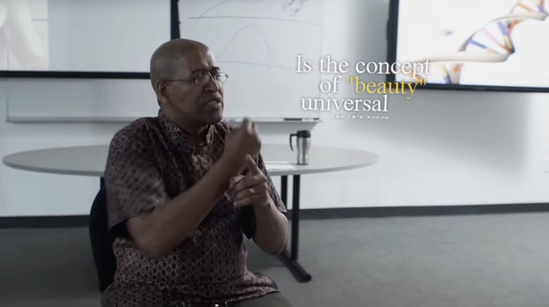 The instructor, seated in a classroom, is signing as subtitles appear next to him on the right side of the screen. The words read, “Is the concept of ‘beauty’ universal." The word "across" is just starting to appear. The sentence is in white text but the word “beauty” appears in yellow for emphasis.