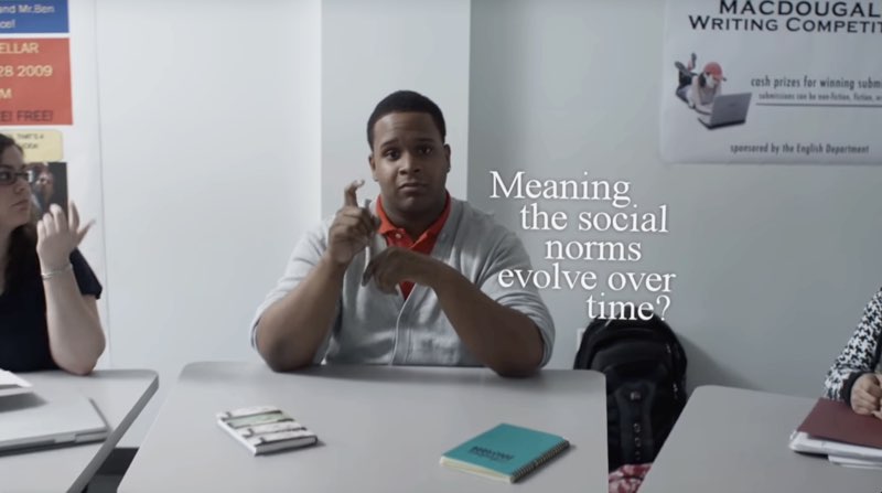 A student is seated at his desk with students at either side. He signs as the white subtitles read, “Meaning the social norms evolve over time?”