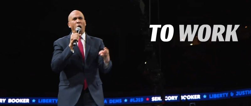 This screenshot of Booker's video shows speaking into a microphone in his hand. White open captions appear that read, "TO WORK,” as they fade onto screen.