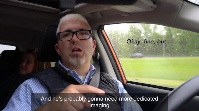 Mick is signing as he drives his car. Open subtitles appear in the car window next to his face that read, "Okay, fine, but..." Closed captions appear at the bottom of the screen saying, "And he's probably gonna need more dedicated imaging."