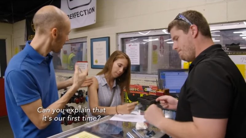 Sheena and Manny are at an indoor firing range with a male employee. Sheena is writing a note as Manny signs to her. Words near Manny read, "Can you explain that it's our first time?"