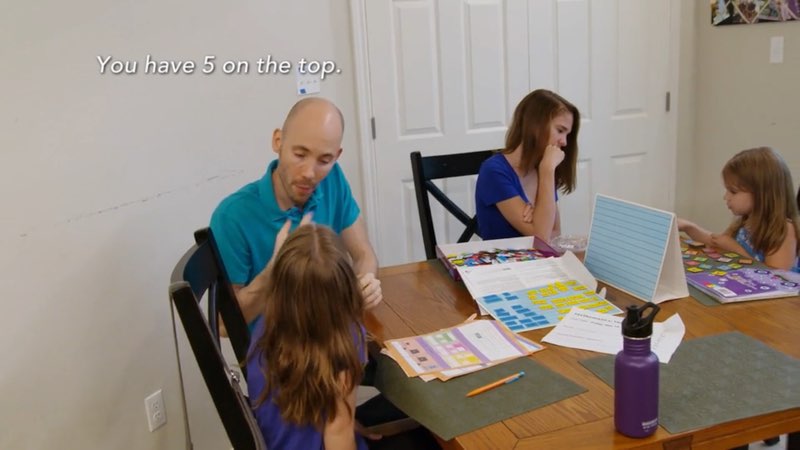 Manny and Sheena are shown at a dining table with two young girls. The children’s math and language activities are on the table. Manny signs to one of his daughters as subtitles appear over his head that read, “You have 5 on the top.”