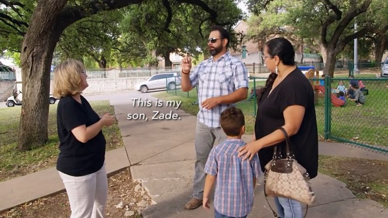 Paco, April, and their son Zade are on a school campus near a playground with another woman from the school. Paco signs to the woman. Open subtitles appear near his lower body close to his son. The open subtitles read, "This is my son, Zade."