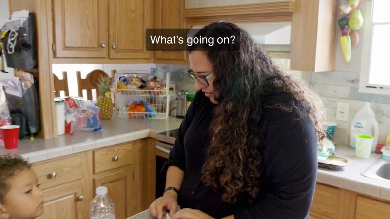 A woman speaks to her toddler son in their kitchen. White closed captions at the top read, "What's going on?"