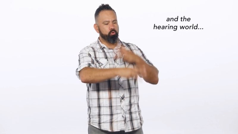 Paco, standing in front of a white background, signs to the camera as he faces his left and signs the word “world.” Open subtitles appear to his left that say, "and the hearing world..."