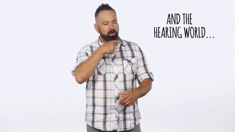 Paco, standing in front of a white background, signs to the camera as he faces his left and signs the word “hearing.” Large, capitalized open subtitles appear to his left that say, "AND THE HEARING WORLD..."