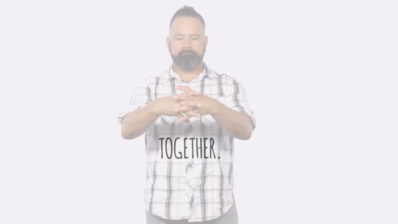 Paco, standing in front of a white background, signs to the camera as he directly faces the camera and signs the word "together."  Large, capitalized open subtitles appear in front of him reading, “TOGETHER.”