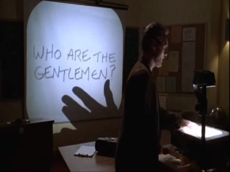 Giles is standing by a projector in a classroom. Words are projected on the screen behind him, in the right direction; they say, “Who are the gentlemen?"