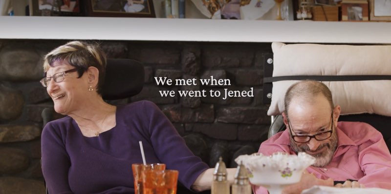Denise and Neil are together in front of a fireplace mantle and holding hands. Open captions appear in the space between them, reading, "We met when we went to Jened."