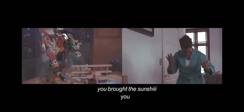 This screen capture of one moment in this music video shows two images side by side. The left image shows children's toys. The right image shows a woman signing the song. There are two rows of captions at the bottom of the screen. The first row reads: "you brought the sunshiii" in italics. The second row reads "you" in regular font.