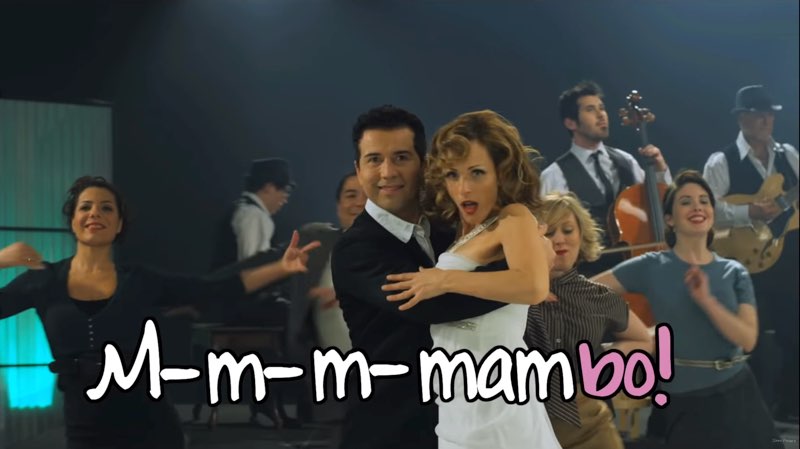 A screenshot from Forbes’ music video show a smiling man and Marlee Matlin dancing the mambo while facing the camera. Musicians and other dancers are in the background. The words on screen read “M-m-m-mambo!” in a large bubbled font, the first four syllables in white and the “bo!” in pink to emphasize the syllable being sung at the moment.