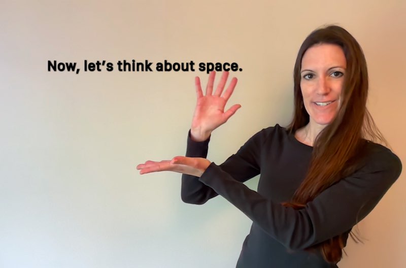 Janine Butler faces the camera while signing “space.” Subtitles appear next to her hands, one of her hands under the subtitles that read, "Now, let's think about space."