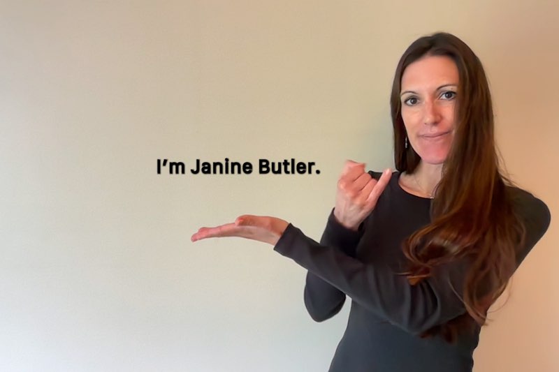 Janine Butler faces the camera as she fingerspells “J.” Subtitles appear next to her hands, with one of her hands under the subtitles. The subtitles read, "I'm Janine Butler."