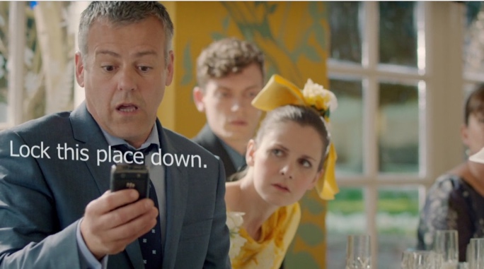 Lestrade is at a wedding in front of a bridesmaid. He is staring at his phone open-mouthed, a startled look on his face. He has straightened up more than in the previous screenshot, so the phone is now closer to his face. The visual text appears across his body between his face and the phone, reading, "Lock this place down."