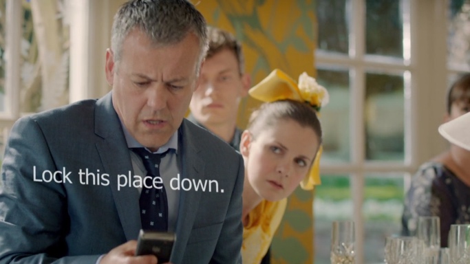 Lestrade is at a wedding in front of a bridesmaid. He is staring at his phone open-mouthed, a startled look on his face. The visual text appears across his body between his face and the phone, reading, "Lock this place down."