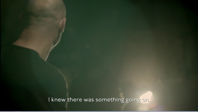 A man faces away from the camera, only the back of his head and shoulders shown. He is in a dark room with a bright lamp at the bottom right of the frame. Subtitles at the bottom read, "I knew there was something going on."