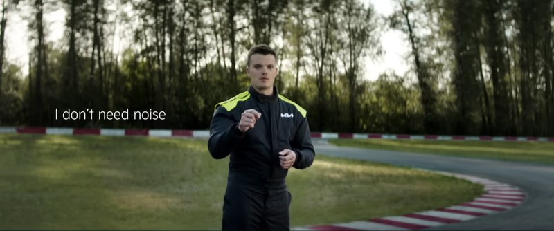 Kris Martin is wearing a black and green Kia race suit by a racetrack. He is signing to the camera. Subtitles appear to his right, and the subtitles read, "I don't need noise."