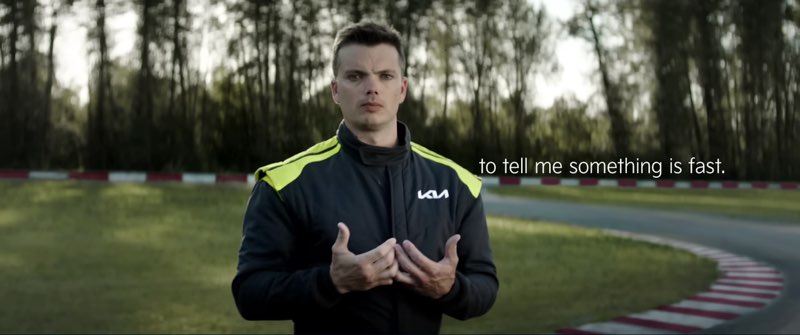Kris Martin iswearing a black and green Kia race suit by a racetrack. He is signing to the camera. Subtitles appear to his left, and the subtitles read, "to tell me something is fast."