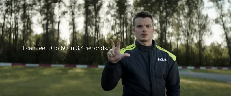 Kris Martin is wearing a black and green Kia race suit by a racetrack. He is signing the number 3. Subtitles appear to his right, and the subtitles read, "I can feel 0 to 60 in 3.4 seconds."